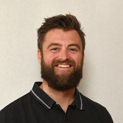 En munter skjeggete mann ved navn Kristian Gilbert, med et varmt smil, iført svart skjorte med krage mot ensfarget bakgrunn.