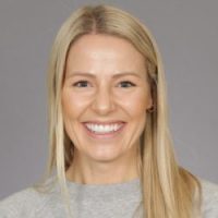 En munter kvinne med langt blondt hår, iført en lys grå topp, smilende til kameraet mot en nøytral grå bakgrunn.