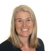 En smilende kvinne med langt blondt hår, iført en mørk topp, mot hvit bakgrunn er Linda M Næsheim.