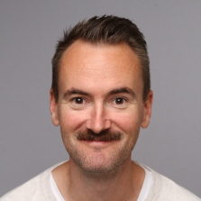 En blid Olav Veland Thu med kort skjegg og bart, smilende til kamera mot en grå bakgrunn.