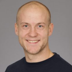 Et portrett av Marius Dahl, en smilende skallet mann med blå øyne, iført en mørkeblå skjorte mot en grå bakgrunn.