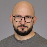 Et portrett av Antonio Egri med skallet hode, iført svarte briller og skjegg, kledd i en uformell grå skjorte mot en nøytral bakgrunn.