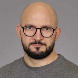 Et portrett av Antonio Egri med skallet hode, iført svarte briller og skjegg, kledd i en uformell grå skjorte mot en nøytral bakgrunn.