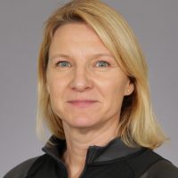 Et profesjonelt portrett av Bente Stensrud med blondt hår, lett smil, iført svart topp og små øredobber, satt mot en grå bakgrunn.