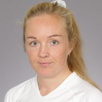 En kvinne, Anne Jorunn Tysseland, med lyst hår bakbundet, klare blå øyne, og et nøytralt uttrykk mot en grå bakgrunn.