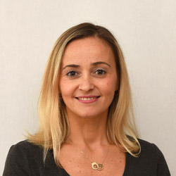 Et profesjonelt hodebilde av Reyhan Caliskan, en smilende kvinne med langt blondt hår, iført en mørk topp og et halskjede med et sirkulært anheng.