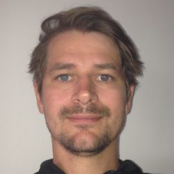 Et portrett av Hans Anton Bjørndalen med rufsete hår, en lys skjeggstubber og et subtilt smil, iført svart skjorte og stående mot en vanlig bakgrunn.