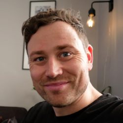 En smilende person med kort hår og en tilfeldig svart t-skjorte, tilsynelatende i koselige innendørs omgivelser med varmt lys, vakkert fanget av fotograf Stian Andersen.