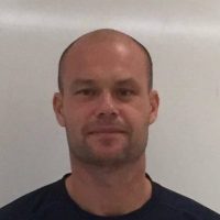 En mann med barbert hode og et innholdsuttrykk iført svart skjorte mot hvit bakgrunn identifiseres som Teemu Pasanen.
