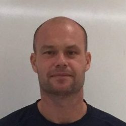 En mann med barbert hode og et innholdsuttrykk iført svart skjorte mot hvit bakgrunn identifiseres som Teemu Pasanen.