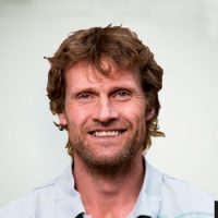 En smilende mann med rufsete brunt hår og skjeggstubber, iført krageskjorte, mot nøytral bakgrunn er Tor Øivind Ødegård.