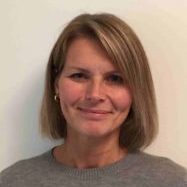 En smilende kvinne med kort blondt hår, iført en grå genser, poserer mot en vanlig hvit bakgrunn.