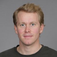 En smilende Andreas Sauge med lyst hår og svart skjorte mot en nøytral grå bakgrunn.