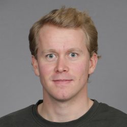 En smilende Andreas Sauge med lyst hår og svart skjorte mot en nøytral grå bakgrunn.