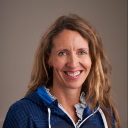 Anne Stine Sånhol, en smilende kvinne med langt, bølget hår iført blå jakke med krageskjorte under, poserer mot en nøytral bakgrunn.