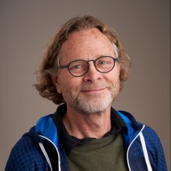 En smilende mann med briller, iført en blå og svart jakke over en grønn skjorte, satt mot en nøytral bakgrunn. Dette er Axel Johan Mellbye.