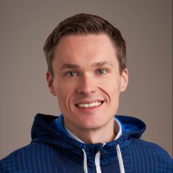 En smilende person med kort hår iført en blåmønstret hettegenser mot en nøytral bakgrunn, fotografert av Bartosz Piasecki.