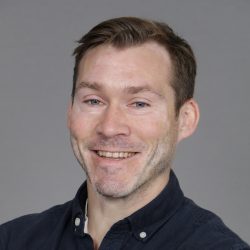 En smilende mann ved navn Carl Ditlev Gran med et vennlig uttrykk, kort hår og lyse skjeggstubber, iført mørk skjorte.