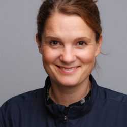 En kvinne med et vennlig smil, iført en mørk bluse og med hestehale mot en nøytral grå bakgrunn.