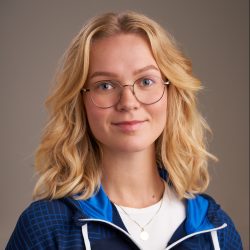 Et portrett av Ellen Westby Agnor, en ung kvinne med blondt, bølget hår og runde briller, iført en blå jakke over en lys topp, satt mot en nøytral bakgrunn. Hun dukker opp