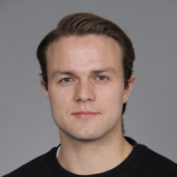 Et portrett av Erik Stuevold med middels langt brunt hår, klar hud og et nøytralt uttrykk som ser direkte på kameraet. Han har på seg en enkel svart skjorte og satt mot en grå bakgrunn