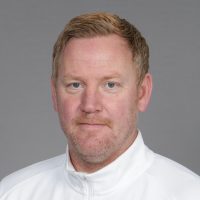 Et portrett av Espen Olsen, med lys hud, sandblondt hår og en fippskjegg, iført hvit topp mot grå bakgrunn. Han ser ut til å ha et gjennomtenkt uttrykk, med en svak