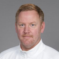 Et portrett av Espen Olsen, med lys hud, sandblondt hår og en fippskjegg, iført hvit topp mot grå bakgrunn. Han ser ut til å ha et gjennomtenkt uttrykk, med en svak