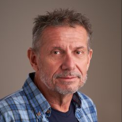 En moden mann ved navn Gunnar Gilde, med salt- og pepperhår og skjeggstubbe, iført blå rutete skjorte, og gir et mildt, ettertenksomt blikk.