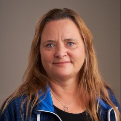 Hanne Espolin Johnson, en kvinne med skulderlangt hår og en blå jakke over en svart skjorte, med et delikat halskjede, ser direkte på kameraet med et nøytralt uttrykk mot en beige bakgrunn.