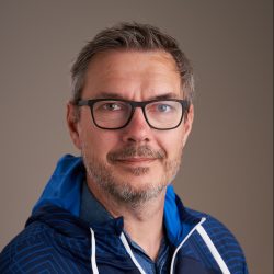 Middelaldrende mann med briller og lys skjeggstubber iført blå jakke, poserer med et selvsikkert smil mot Harald Iuell-bakgrunn.