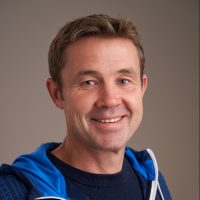 En smilende Harald Lindal med et varmt uttrykk, iført blå sportsjakke med hvite detaljer, satt mot en nøytral grå bakgrunn.