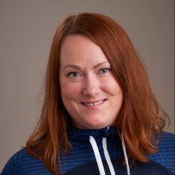 En smilende Ida Fløisand med skulderlangt rødt hår, kledd i en blå sportstopp med hvit snøring, mot nøytral bakgrunn.