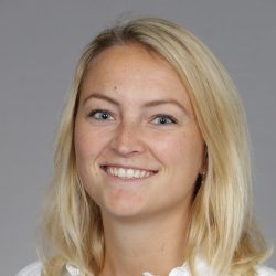 En kvinne med blondt hår og blå øyne som smiler til kameraet mot en grå bakgrunn, kjent som Ine Wigen.
