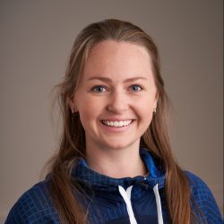 En smilende kvinne med blå øyne og langt blondt hår bundet tilbake, iført en blåmønstret hettegenser med glidelås, poserer for et profesjonelt portrett mot en nøytral bakgrunn.