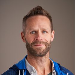 En selvsikker mann med kort skjegg og intenst blikk som poserer for et portrett, iført en blå og grå jakke over en uformell skjorte, som minner om Jørgen Huitfeldts stil.