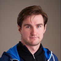 Et portrett av Karl Anders Sletsjøe, en selvsikker mann med brunt hår og blå øyne, iført blå og hvit sportsjakke mot nøytral bakgrunn.
