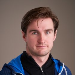 Et portrett av Karl Anders Sletsjøe, en selvsikker mann med brunt hår og blå øyne, iført blå og hvit sportsjakke mot nøytral bakgrunn.