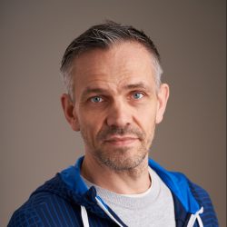 Et portrett av Lars Indresand med skjeggstubber og salt-og-pepper-hår, som gir et lett, ettertenksomt smil til kameraet mot en nøytral bakgrunn. Han har på seg en blå og hvit hettegenser med