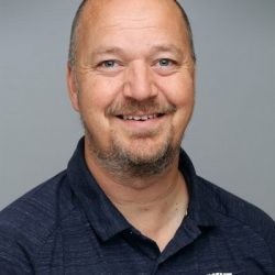 En munter mann med et vennlig smil, skallet på toppen og med kort hår på sidene, iført en mørk skjorte med krage. han fremstår imøtekommende og selvsikker, satt mot en nøytral grå bakgrunn.