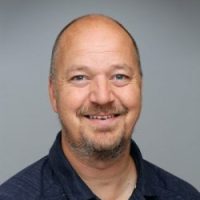 En munter mann med et vennlig smil, skallet på toppen og med kort hår på sidene, iført en mørk skjorte med krage. han fremstår imøtekommende og selvsikker, satt mot en nøytral grå bakgrunn.