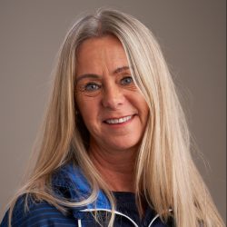 Selvsikker og balansert, Liv Mona Ramberg, en moden kvinne med langt blondt hår og et varmt smil, poserer mot en nøytral bakgrunn, og utstråler en atmosfære av tilgjengelighet og erfaring.