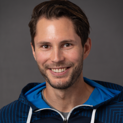 Portrett av Magnus Nilsen, en smilende mann med skjeggstubber, iført en blå hettegenser, som utstråler en vennlig og imøtekommende stemning.