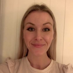 En kvinne med blondt hår, smiler forsiktig til kameraet, i et rom med en lys vegg. Malene Namstad Saxhaug står selvsikkert, uttrykket hennes innbydende og varmt.