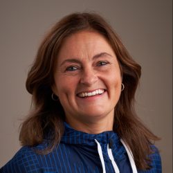 En smilende kvinne med langt brunt hår, iført en blå topp med mønstret design, er Marit Nordhagen.