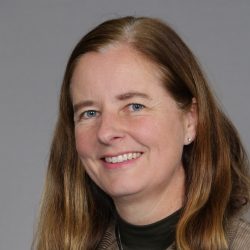 Kvinne med et varmt smil, langt brunt hår, iført brun jakke og mørk turtleneck, mot en nøytral grå bakgrunn er Marit Billington Larsen.