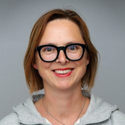 En munter kvinne med skulderlangt hår iført svarte briller og en grå hettegenser, smilende til kameraet mot en nøytral grå bakgrunn.