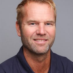 En smilende mann med kort blondt hår, skjeggstubber og et selvsikkert blikk, iført en mørk poloskjorte mot en nøytral bakgrunn.