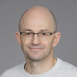 Et portrett av smilende Olivier Graziani, en middelaldrende mann med briller, et tett trimmet fippskjegg og et skallet hode, iført en lys genser med rund hals mot en nøytral grå bakgrunn