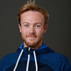 Selvsikker ung mann med et lite smil iført Robin Gruben blå sportsjakke med hvite snorer, satt mot en nøytral grå bakgrunn.