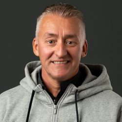 En smilende Øyvind Røsland med kort grått hår iført en tilfeldig grå hettegenser mot mørk bakgrunn.